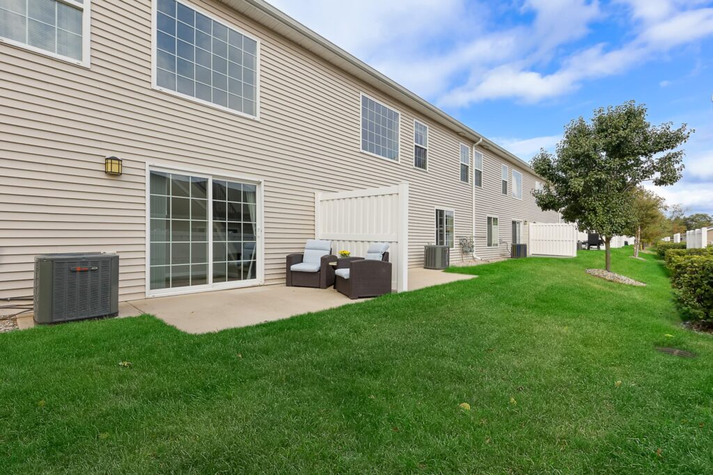Two-story beige townhouse with large windows, a concrete patio, outdoor seating, air conditioning units, and a grassy yard bordered by white fencing and small trees.