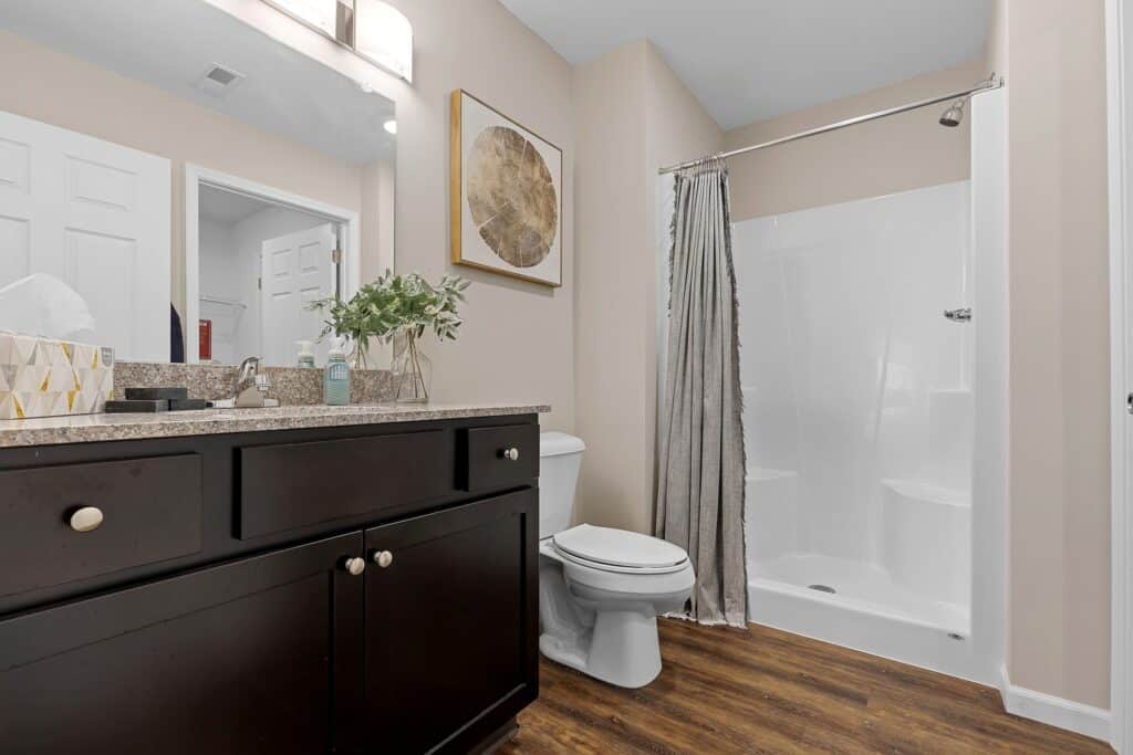 Modern bathroom with dark wood vanity, white countertop, toilet, shower with curtain, wall art, and wood-style flooring.