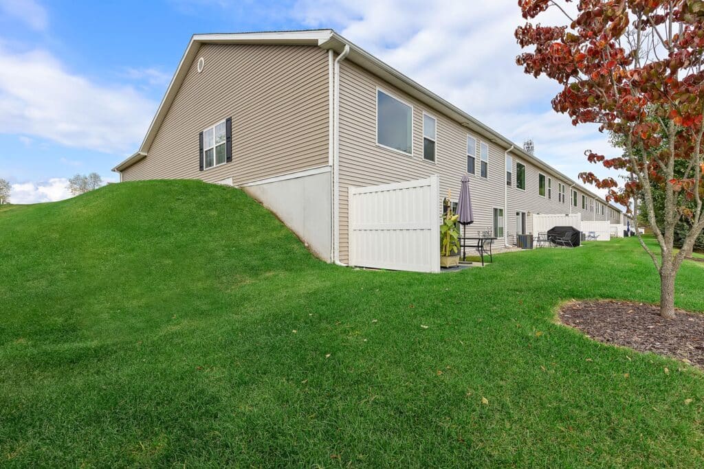 Row of tan townhouses with small patios and a white privacy fence, surrounded by green grass and a tree with red leaves under a partly cloudy sky.
