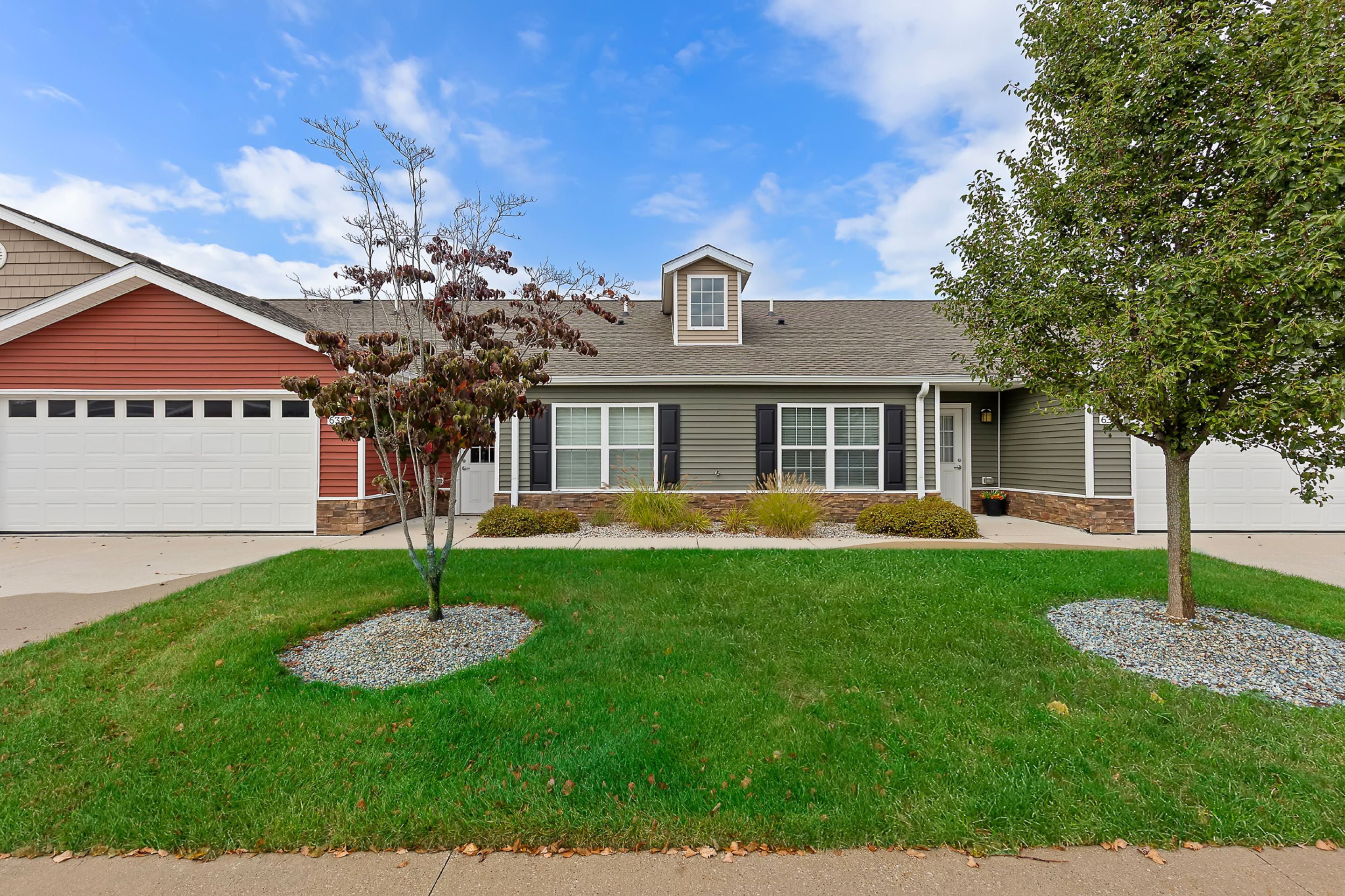 Single-story suburban house with gray siding, white trim, and attached garage, flanked by two trees and a well-kept lawn under a partly cloudy sky.