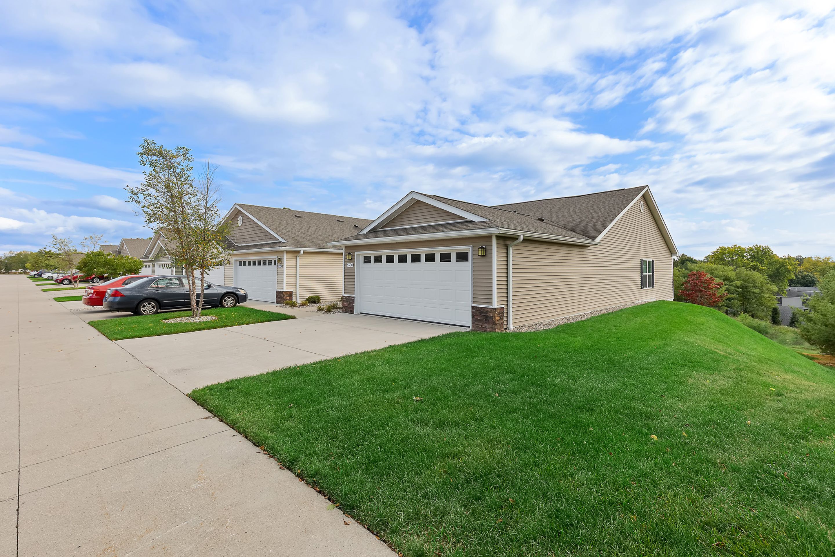 Single-story suburban houses with attached garages, a driveway, and well-maintained lawns under a partly cloudy sky. Several cars are parked along the street.