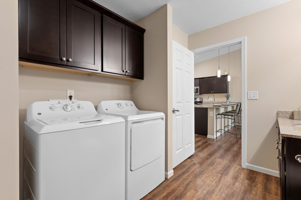 Laundry area with a white washer and dryer, dark upper cabinets, and wood flooring, adjacent to a kitchen visible through an open door.