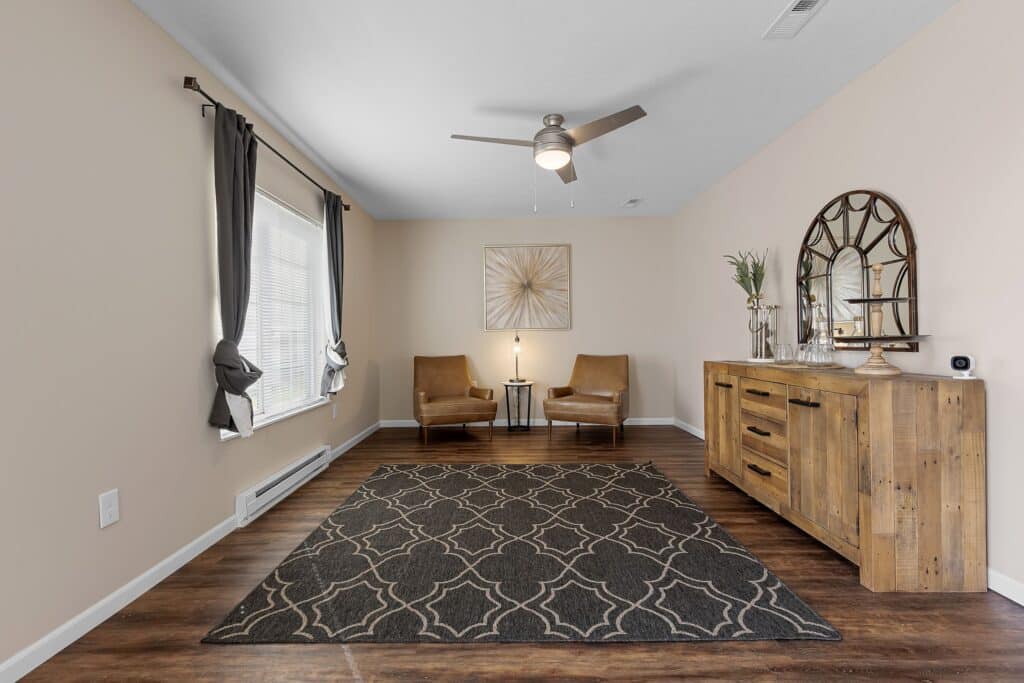 A living room with two brown chairs, a small round table, a patterned area rug, a wooden sideboard with decor, and a ceiling fan. Neutral walls and dark curtains frame a window.