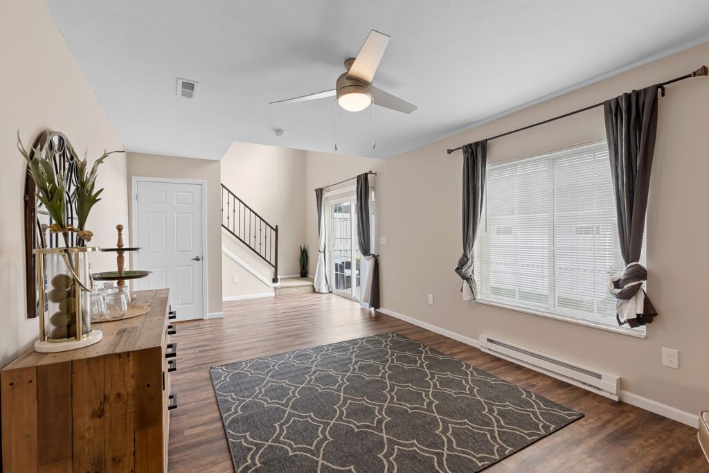 A living room with a ceiling fan, patterned rug, large window with curtains, wood flooring, and a wooden console table; stairs and a door are visible in the background.