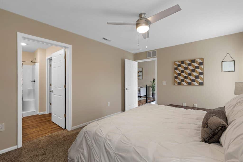 Bedroom with beige walls, a bed with white bedding, a ceiling fan, and open doors leading to a bathroom and hallway. Geometric artwork hangs above the bed.