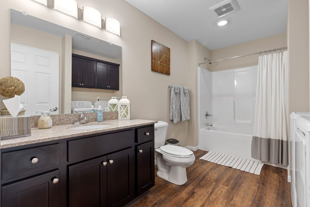 Modern bathroom with dark wood cabinets, granite countertop, white toilet, shower-tub combo with curtain, and wood-look flooring. Neutral decor and wall-mounted lights above the mirror.