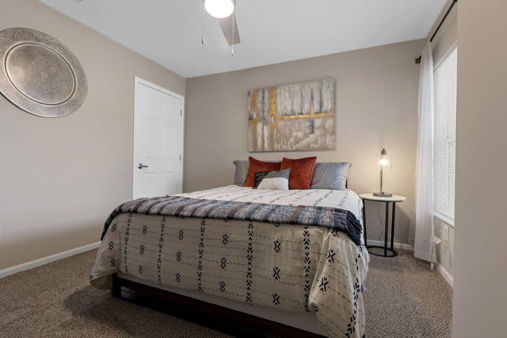 A neatly made bed with patterned bedding and pillows sits in a neutral-toned bedroom with carpet, a round wall decor, abstract art, a nightstand, and a lamp by the window.