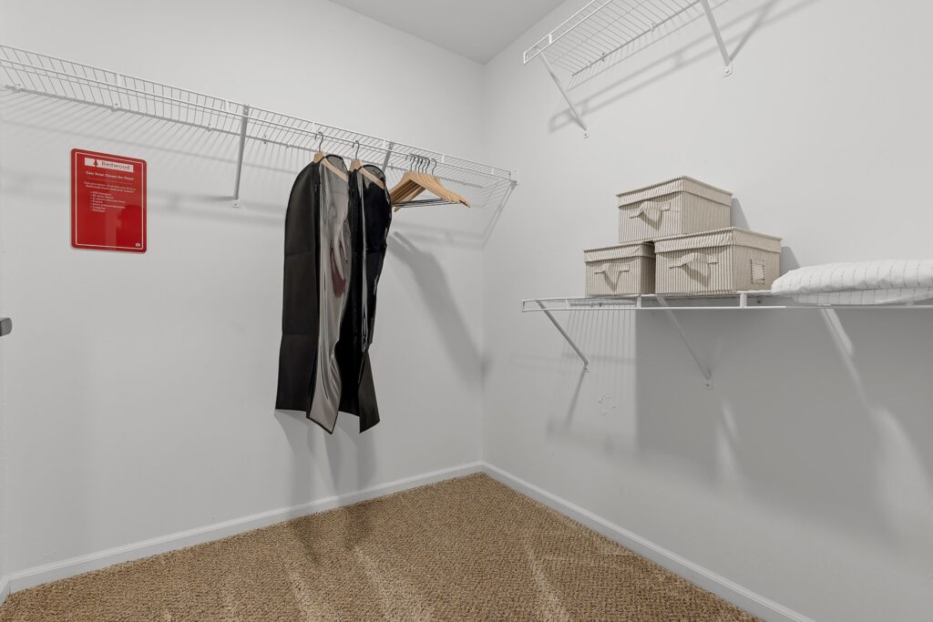 A walk-in closet with wire shelves, a few hanging clothes, three storage boxes, a pillow, and a red notice on the wall. The floor is carpeted.