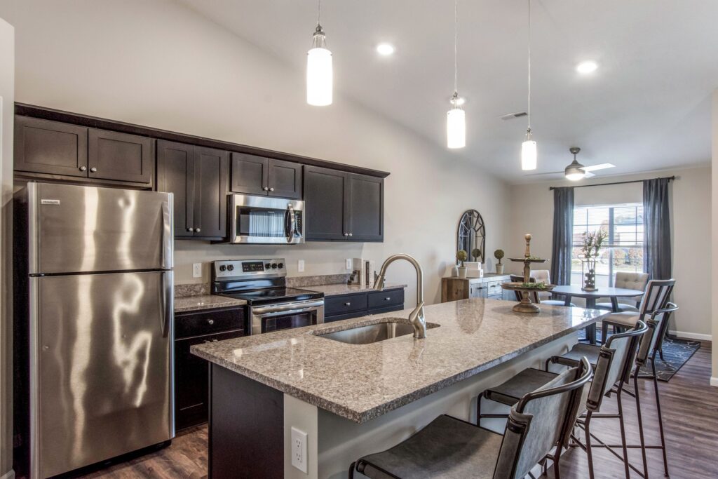 Modern kitchen with dark cabinets, stainless steel appliances, a large granite island with sink, and bar stools; dining area and living room visible in the background.