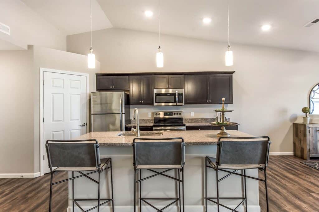 Modern kitchen with a granite island, three barstools, stainless steel appliances, dark cabinets, and pendant lighting.
