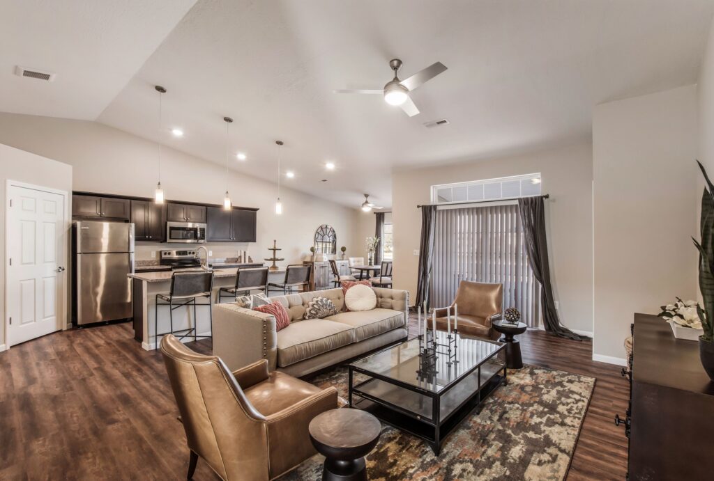 Open-concept living area with modern kitchen, stainless steel appliances, dining table, and a cozy seating arrangement on wood flooring, lit by ceiling lights and natural light through glass doors.