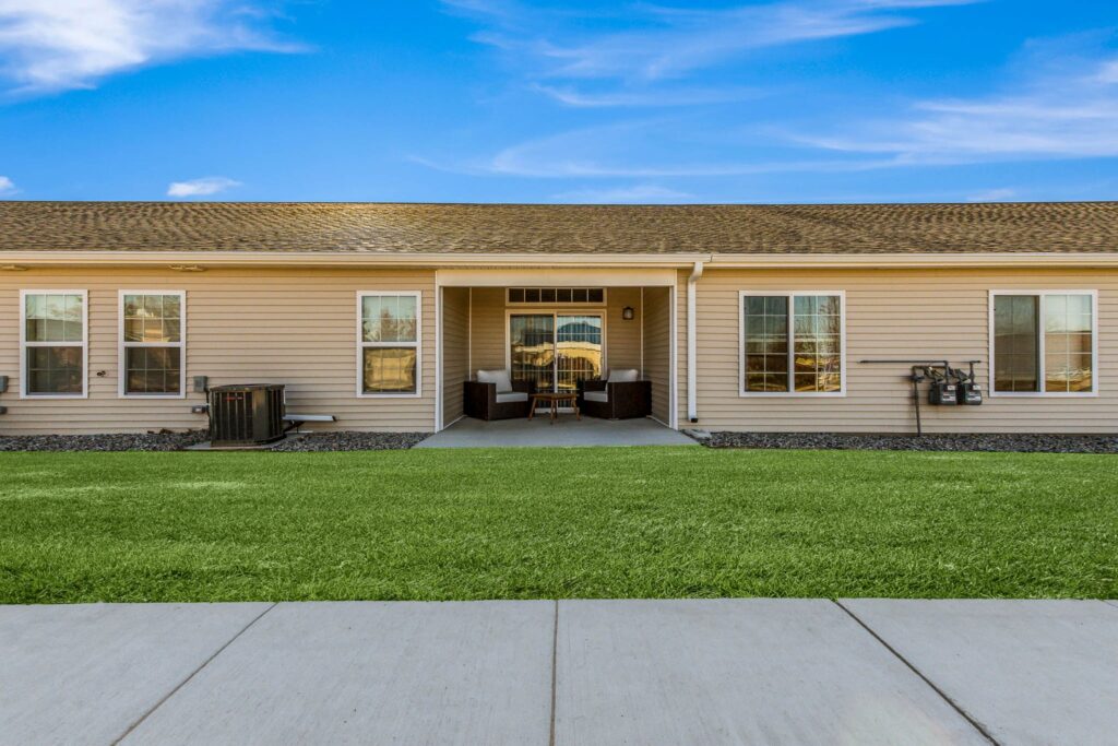 Single-story beige house with a covered patio, multiple windows, and well-kept green lawn under a blue sky with scattered clouds.