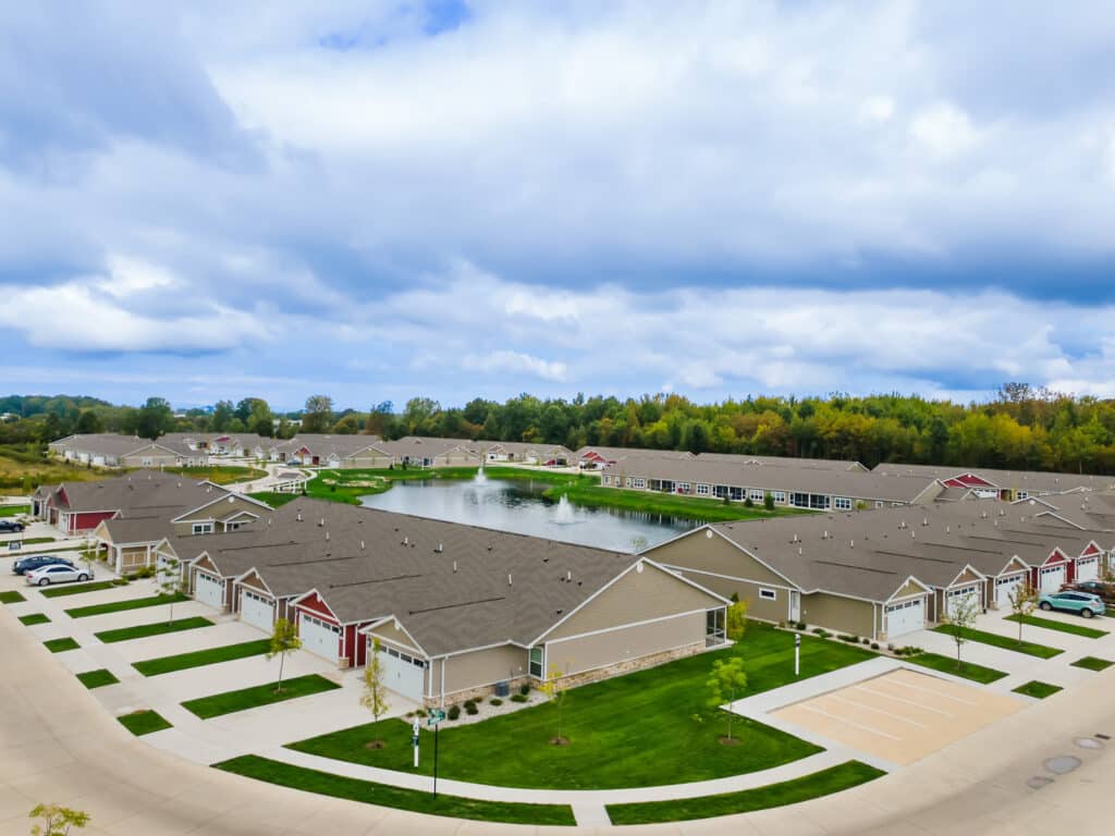 Aerial view of a suburban residential community with rows of single-story houses, parking lots, and a central pond surrounded by trees under a cloudy sky.