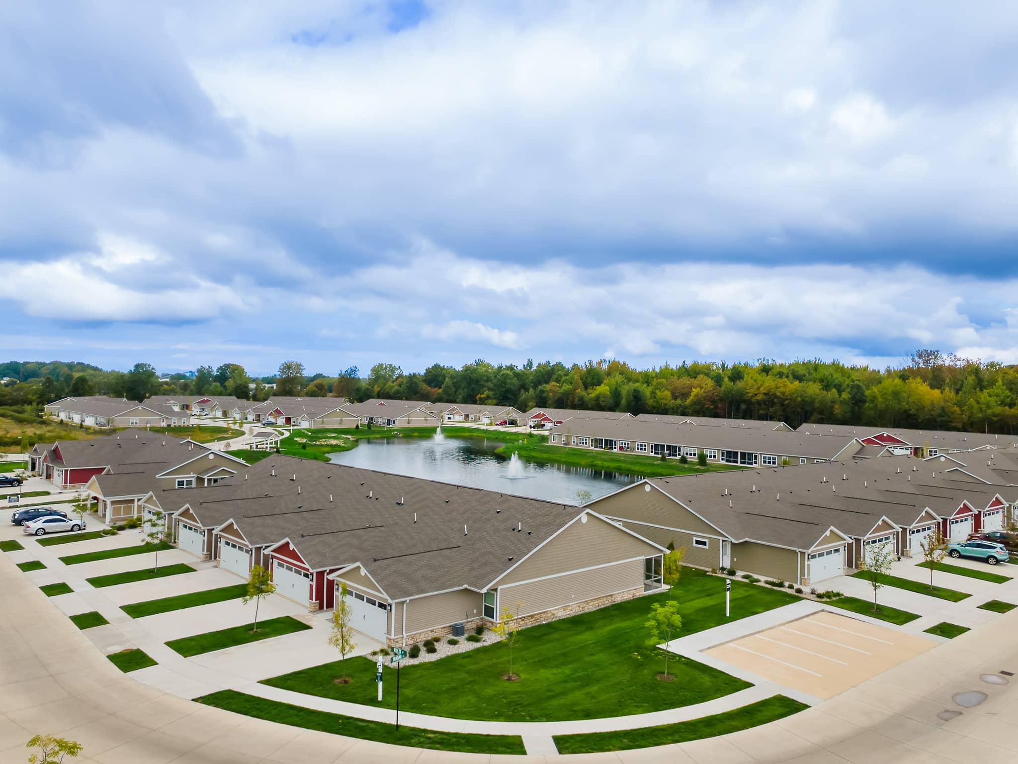 Aerial view of a suburban residential community with rows of single-story houses, parking lots, and a central pond surrounded by trees under a cloudy sky.