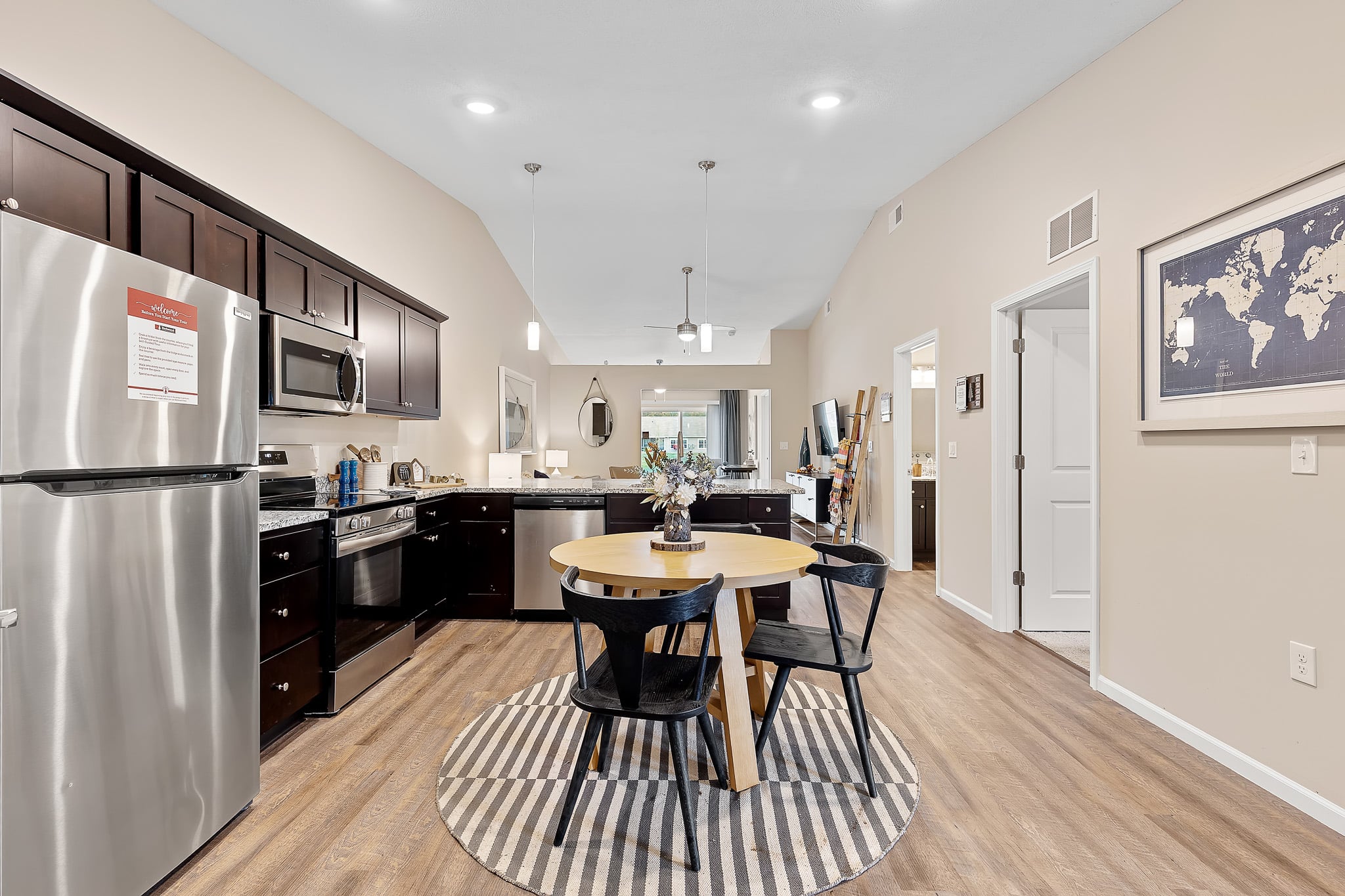 Modern kitchen and dining area with dark wood cabinets, stainless steel appliances, a round wooden table with four black chairs, and a striped rug on a wooden floor.