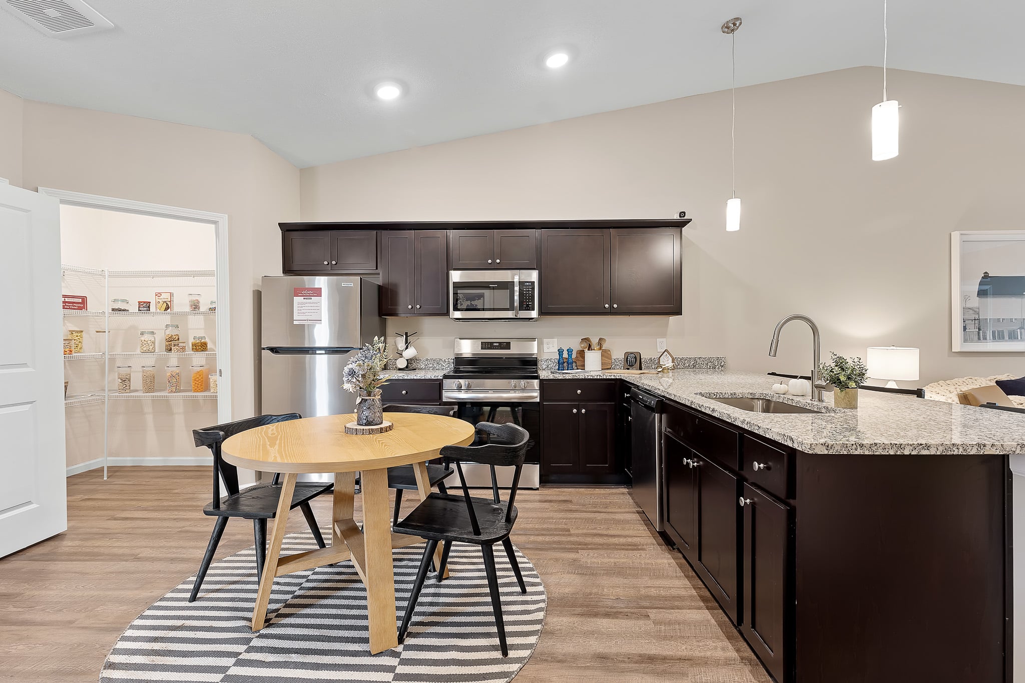 Modern kitchen with dark cabinets, stainless steel appliances, granite countertops, and a small round dining table with four chairs on a striped rug. Open pantry is visible in the background.