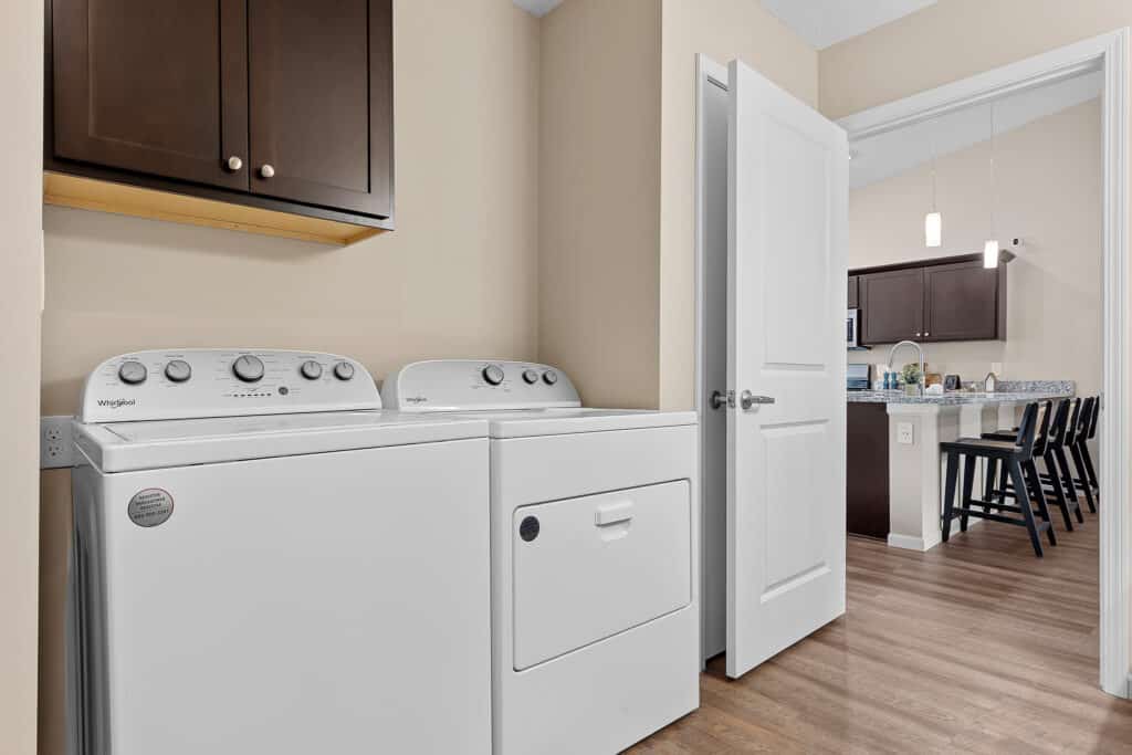 Laundry room with a white washer and dryer beneath dark cabinets, adjacent to an open door leading to a kitchen with bar stools and pendant lights.