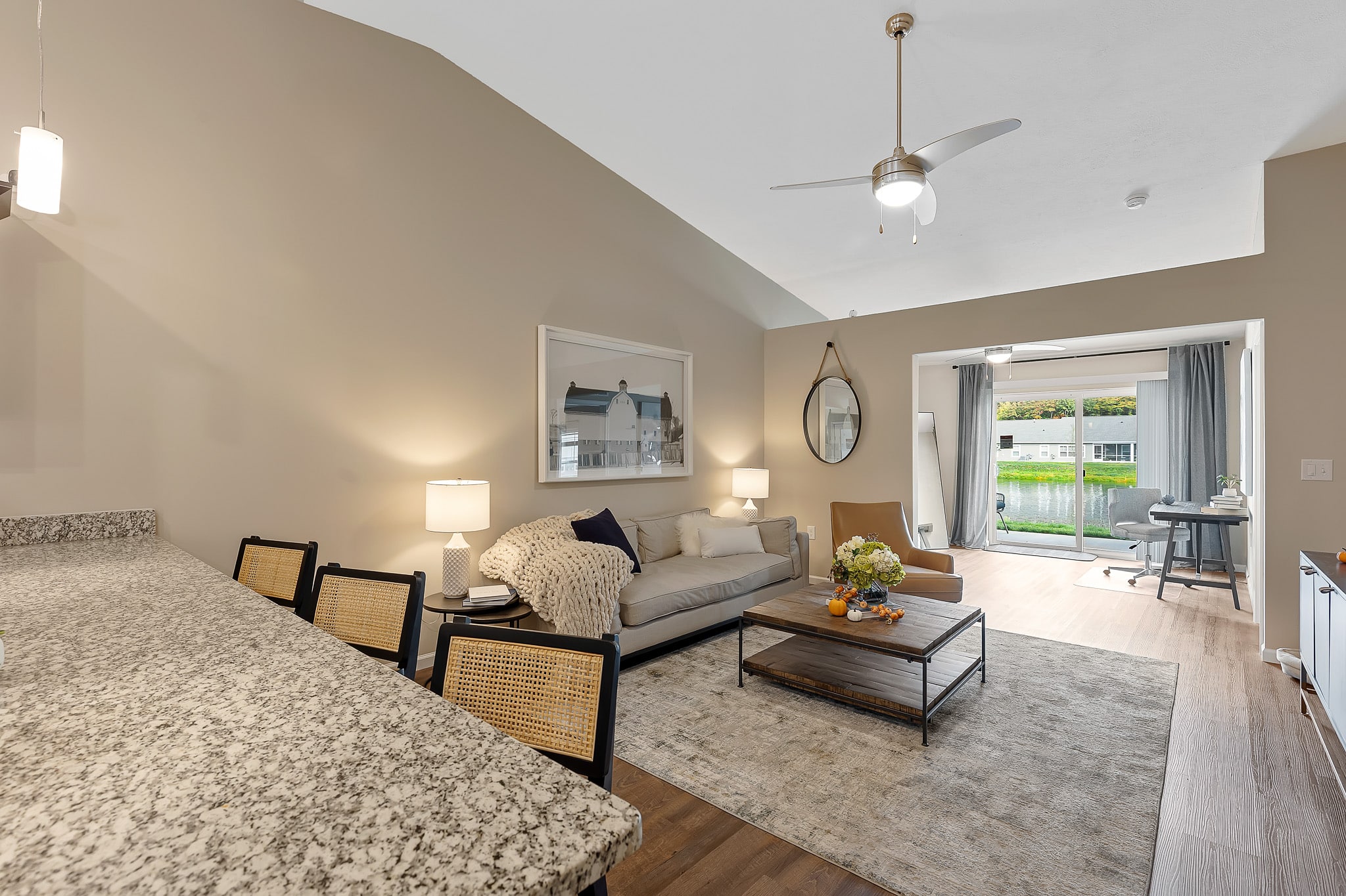 Open-concept living room with a sofa, armchair, coffee table, and large window; kitchen counter with bar stools in foreground; neutral tones and modern decor.