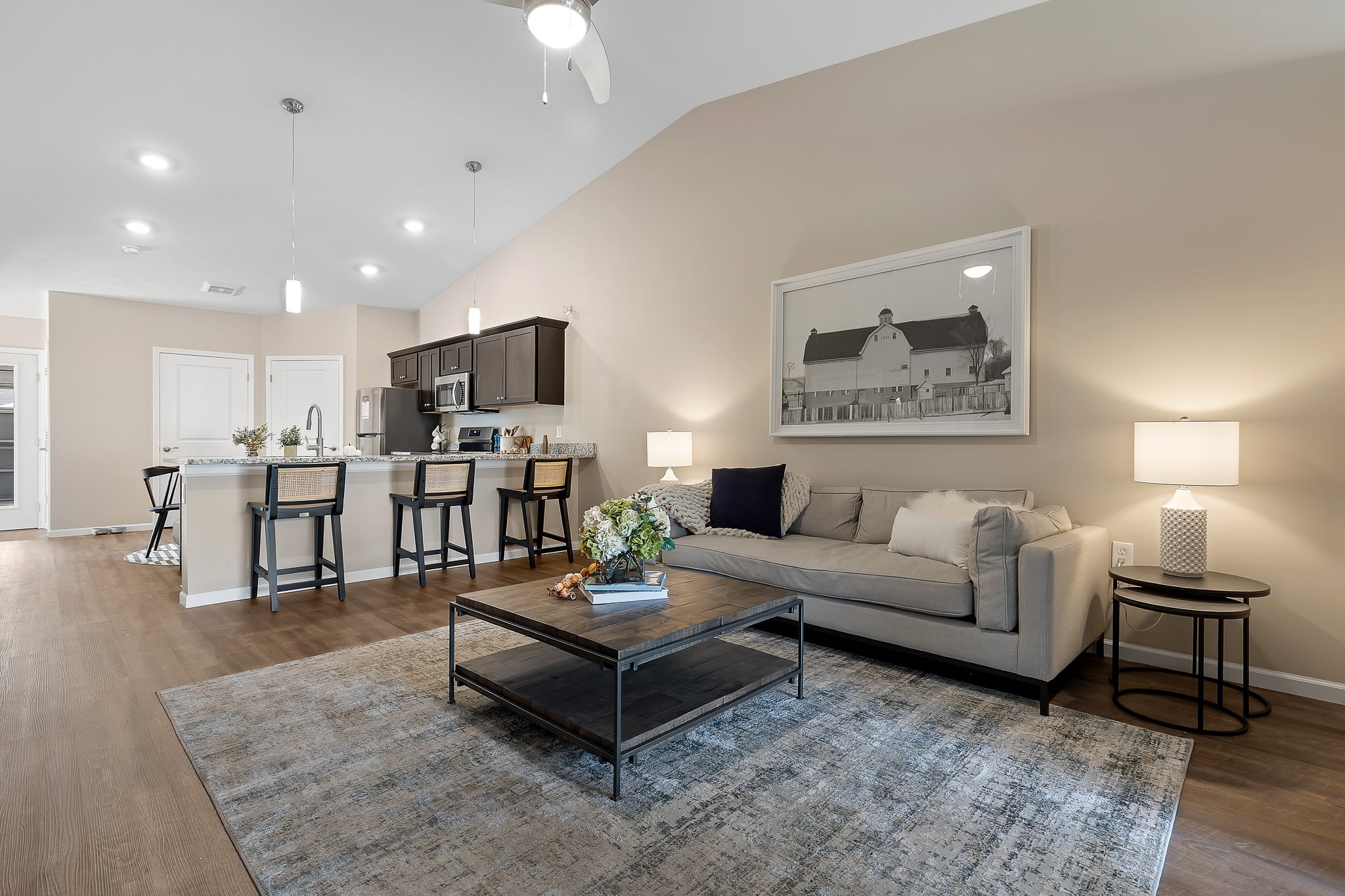 Modern open-concept living room and kitchen with neutral tones, a gray sofa, coffee table, bar stools, and pendant lighting; large barn artwork hangs on the wall above the sofa.