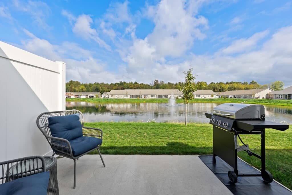 Patio with two chairs and a gas grill overlooks a pond with a fountain, surrounded by grass and residential buildings under a partly cloudy sky.