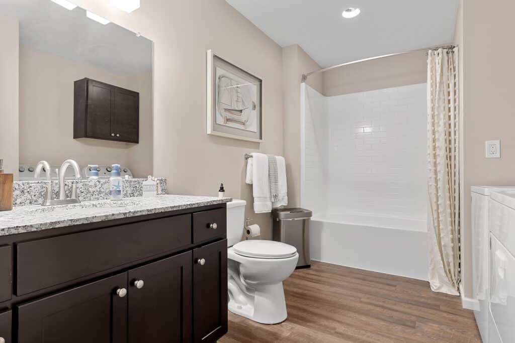 Modern bathroom with a granite countertop vanity, wall mirror, toilet, white bathtub with tiled shower, wood-look flooring, and neutral-colored walls.
