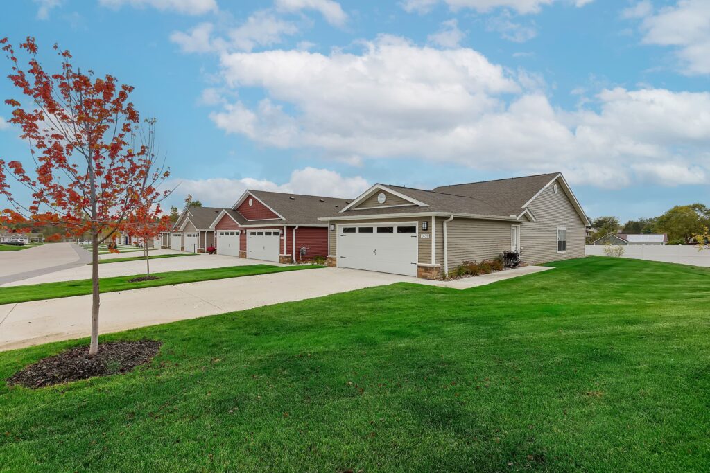 Row of single-story suburban houses with attached garages, green lawns, a sidewalk, and a young tree with red leaves under a partly cloudy sky.