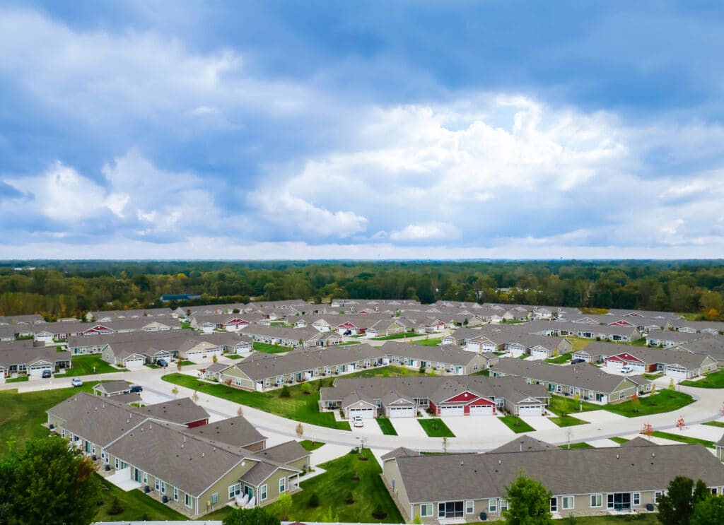 Aerial view of a suburban neighborhood with rows of identical houses, green lawns, and curving streets under a partly cloudy sky.