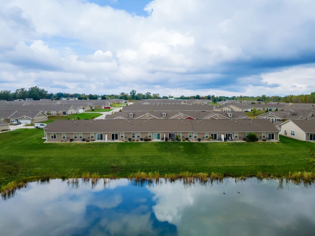A row of single-story houses with backyards faces a small pond reflecting the cloudy sky in a suburban neighborhood.