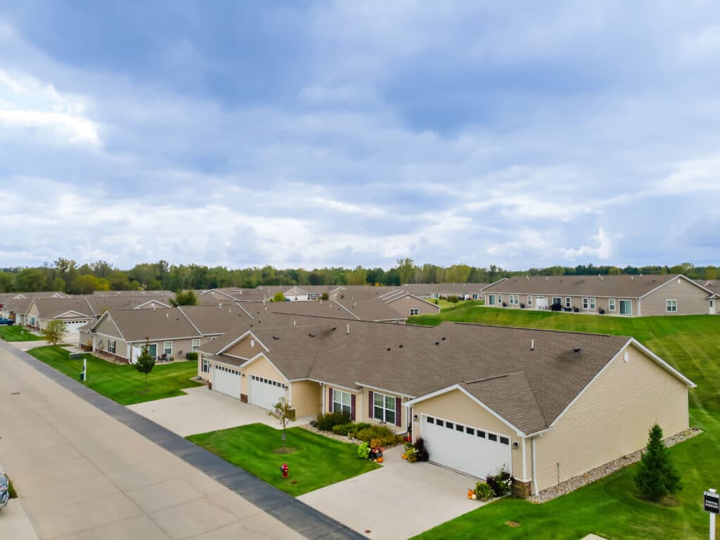 A suburban neighborhood with single-story houses, neatly manicured lawns, and a wide paved street under a cloudy sky.