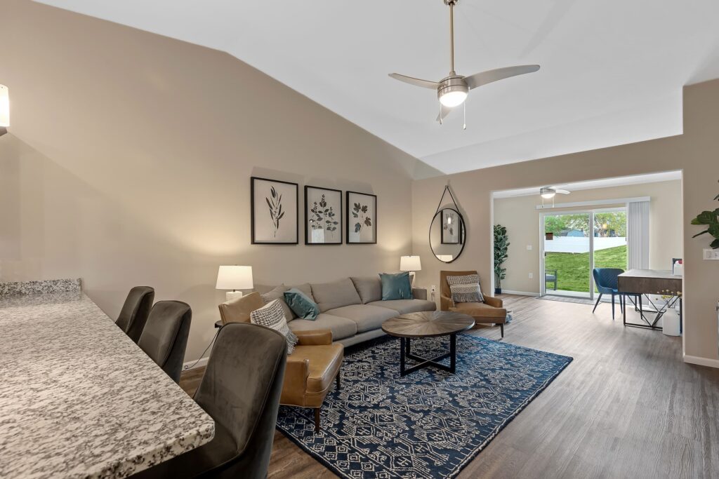 Modern living room with a gray sofa, blue rug, and tan chairs, adjacent to a breakfast bar and sliding glass doors leading to a grassy outdoor area.
