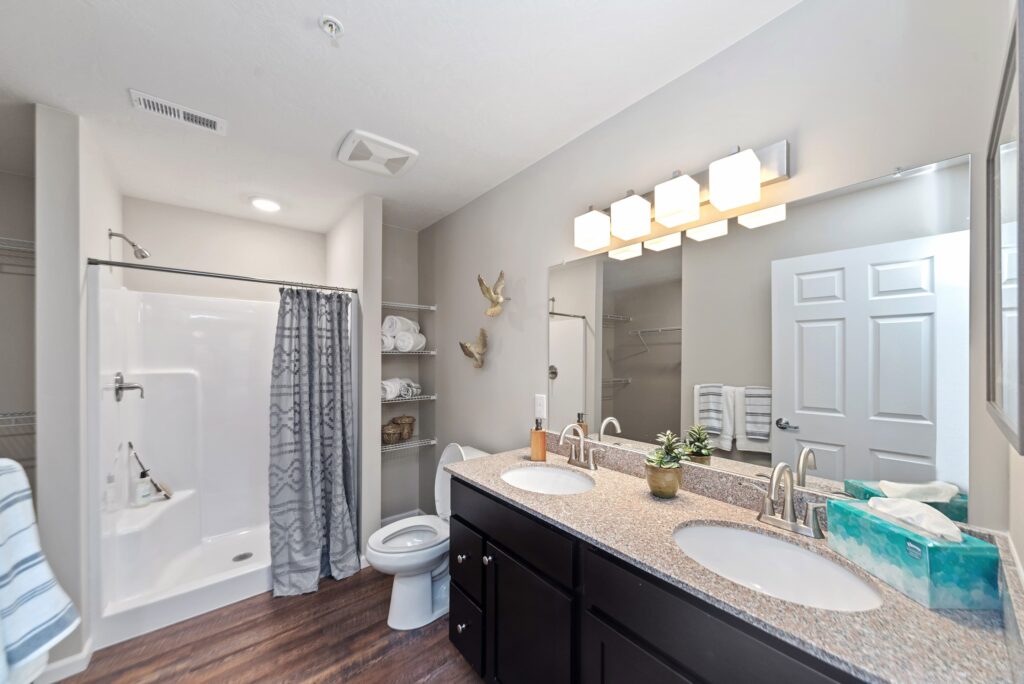 Modern bathroom with a double-sink vanity, granite countertop, large mirror, shower with patterned curtain, toilet, shelves with towels, and wood-look flooring.