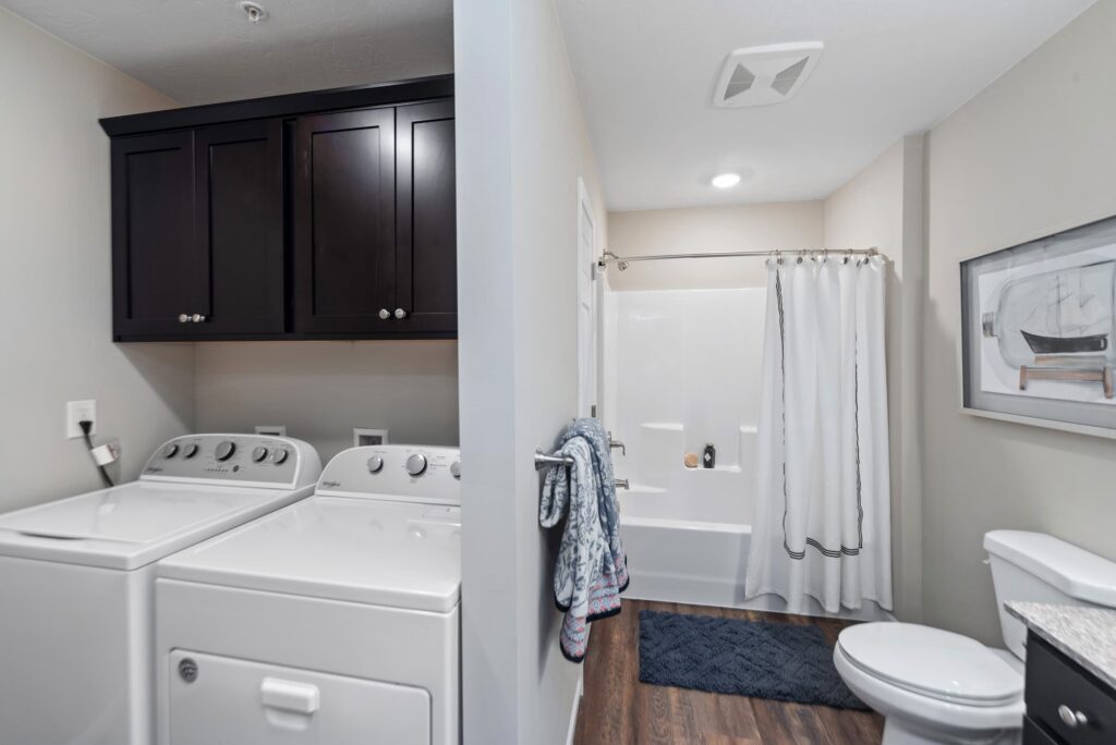 A laundry area with a washer and dryer is adjacent to a bathroom with a bathtub, toilet, sink, wall art, and a dark wood floor.