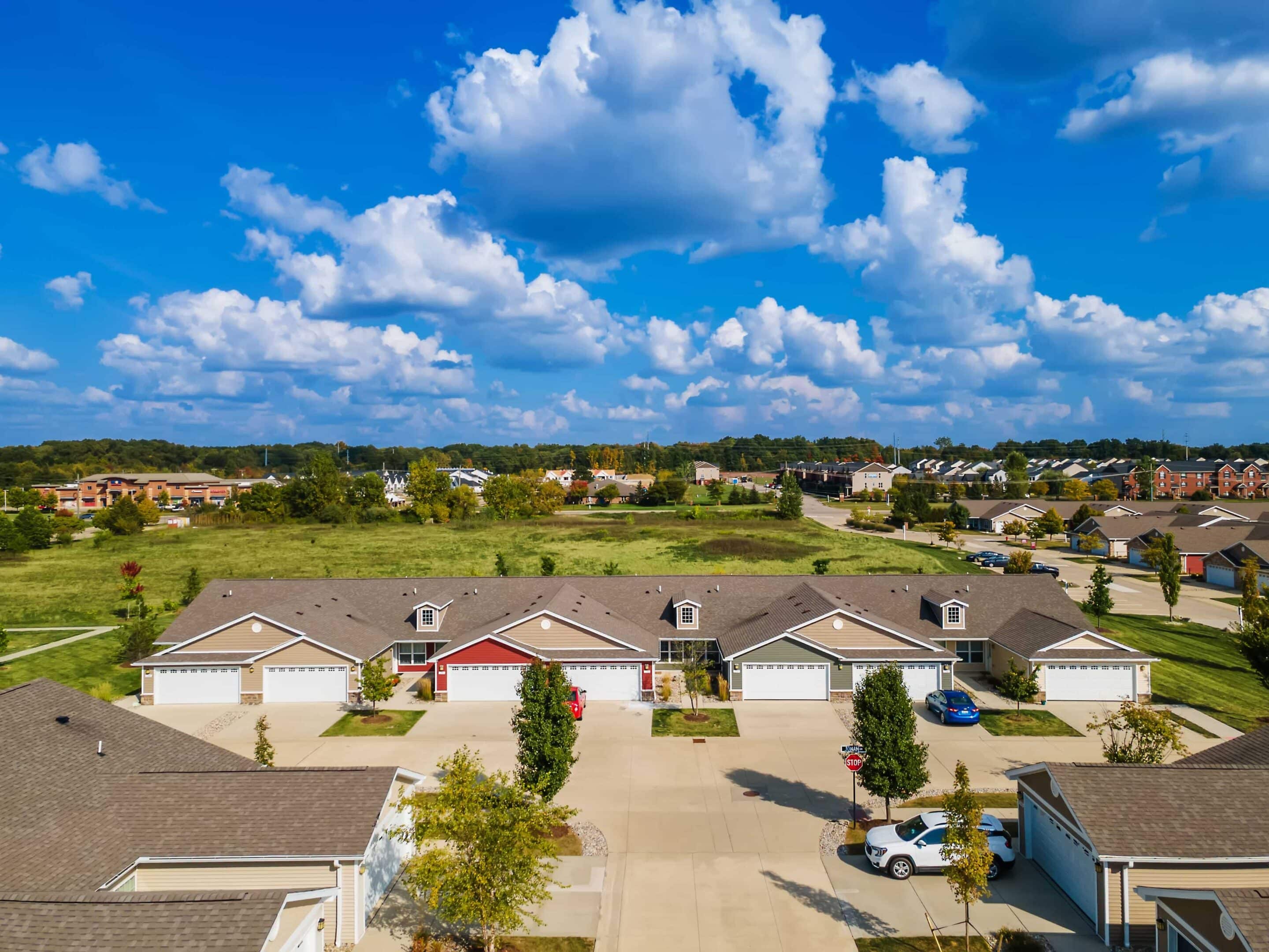 Aerial view of a suburban residential area featuring rows of single-story houses with garages, driveways, and green spaces under a partly cloudy sky.