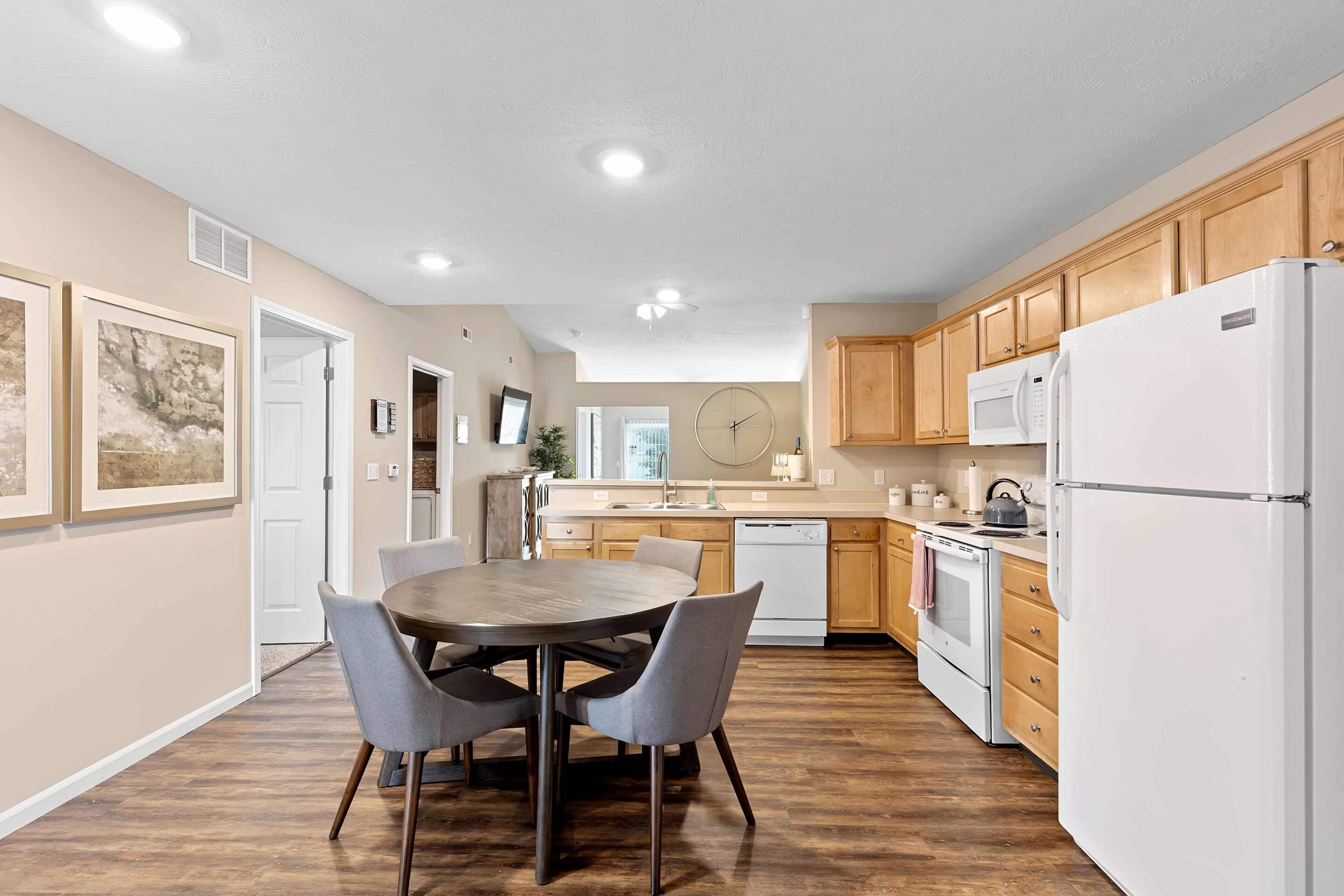 A modern kitchen with wood cabinets, white appliances, a round dining table with four gray chairs, and wall art, featuring wood floors and neutral walls.