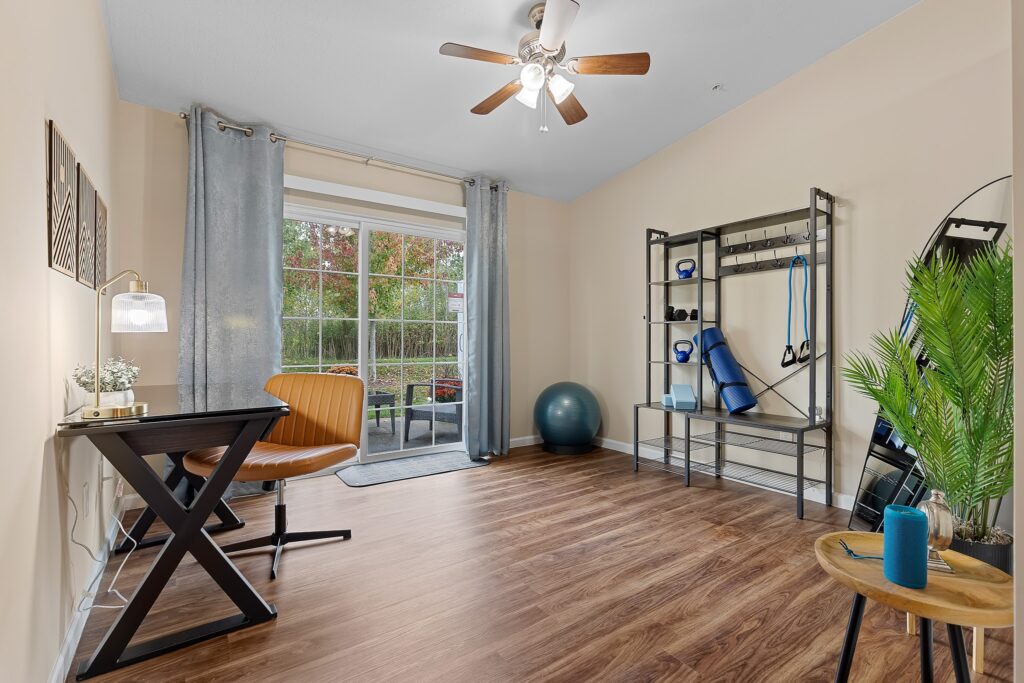 A home office and workout space with a desk, chair, indoor plant, exercise equipment, and large window with outdoor view.