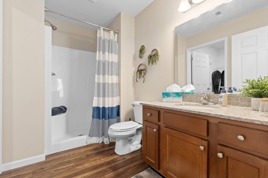 Modern bathroom with a striped shower curtain, toilet, wooden vanity, large mirror, and wall-mounted plant decor. The floor is wood-style and countertop holds soap and tissues.