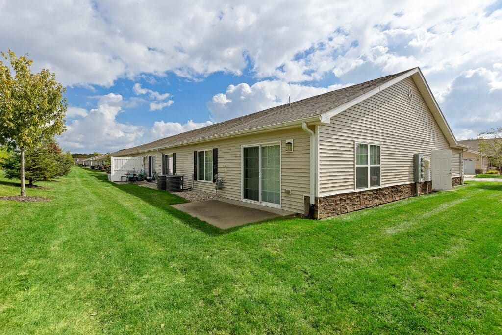 Single-story beige house with sliding glass doors, a small concrete patio, and a well-maintained lawn under a partly cloudy sky.
