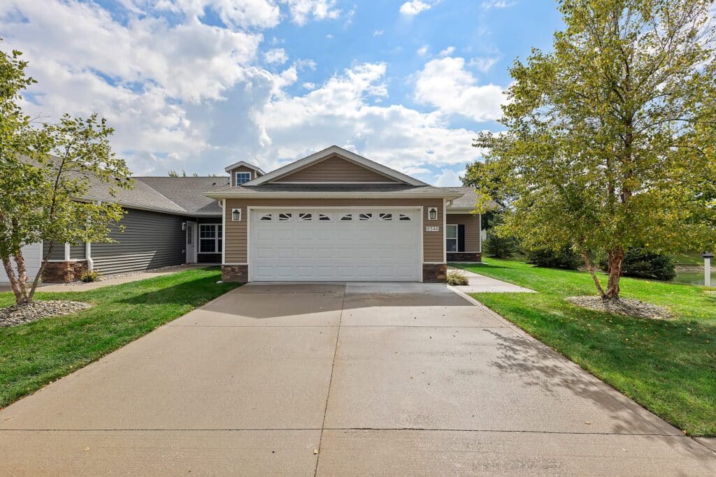 Single-story suburban house with a double garage, beige siding, and a concrete driveway, surrounded by green grass, trees, and a partly cloudy sky.