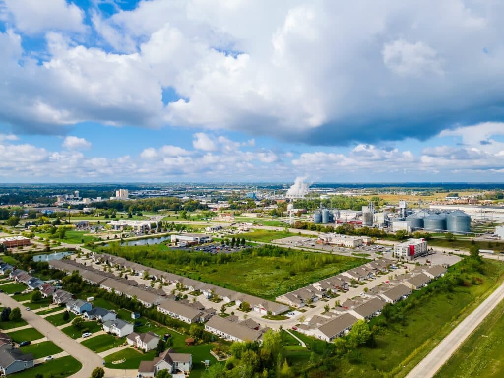 Aerial view of a suburban neighborhood with houses, green spaces, and nearby industrial buildings under a partly cloudy sky.
