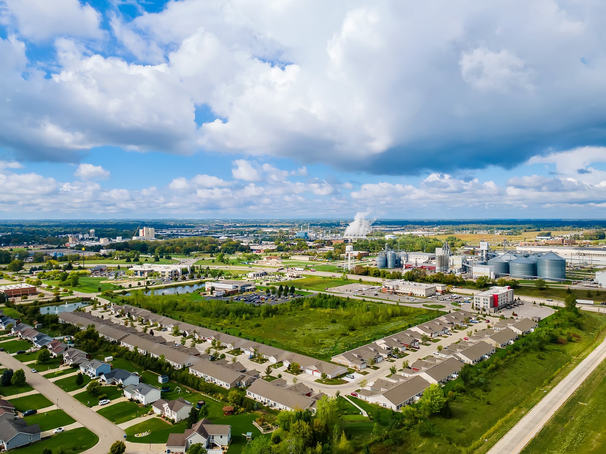 Aerial view of a suburban neighborhood with houses, green spaces, and nearby industrial buildings under a partly cloudy sky.