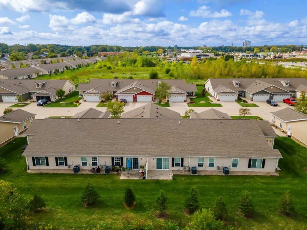 Aerial view of a suburban neighborhood with single-story houses, driveways, green lawns, and rows of trees under a partly cloudy sky.