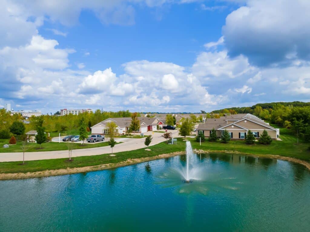 A small pond with a fountain in front of single-story houses and a parking area, surrounded by trees under a partly cloudy sky.