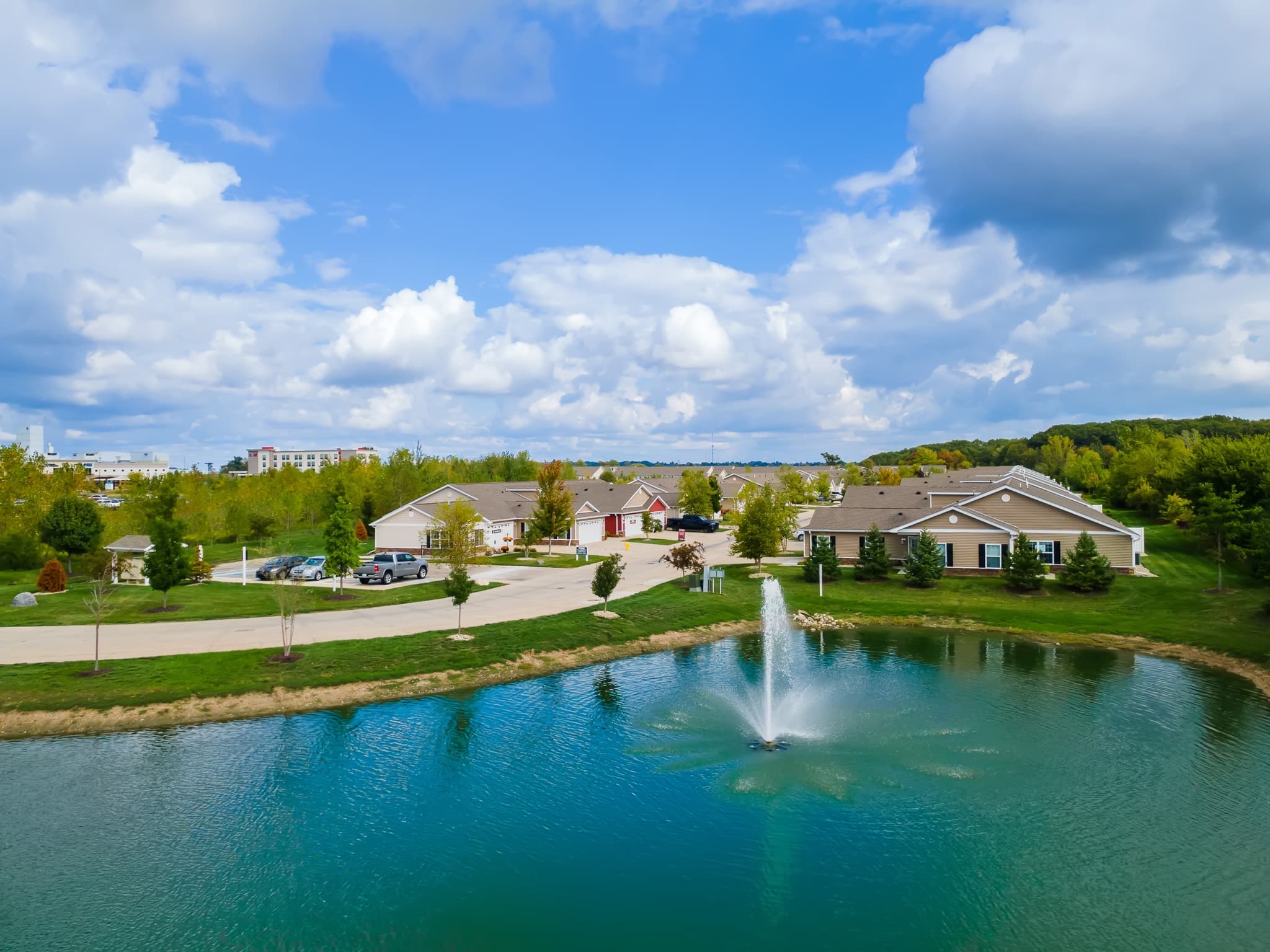 A small pond with a fountain in front of single-story houses and a parking area, surrounded by trees under a partly cloudy sky.