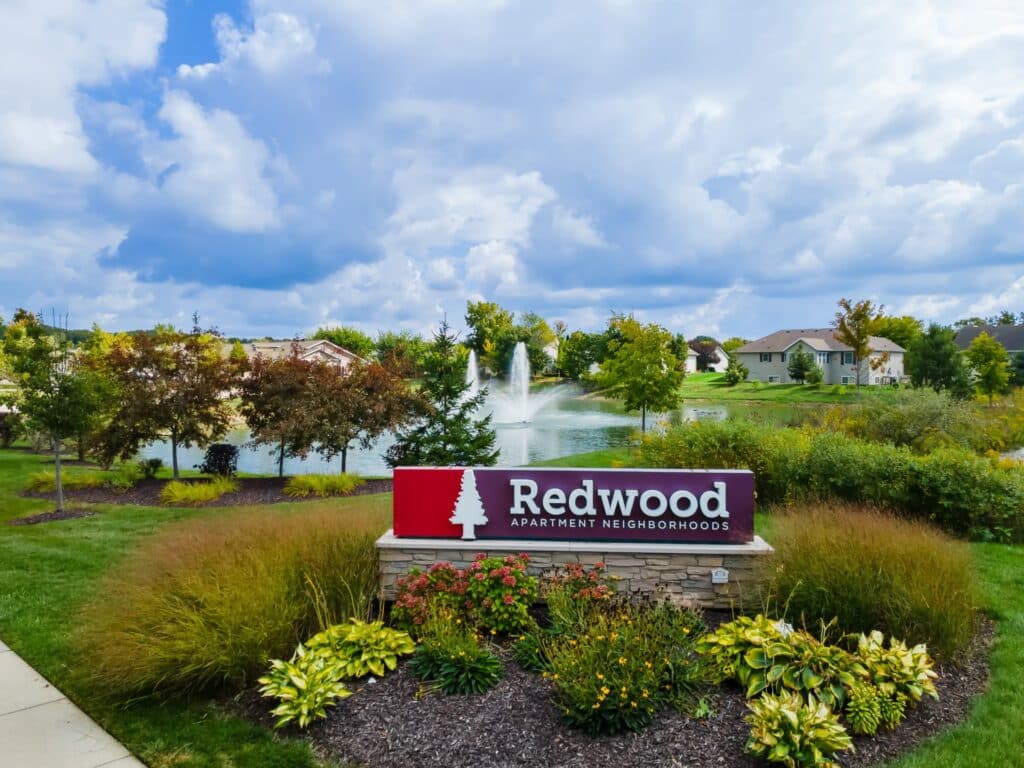 A landscaped entrance sign for Redwood Apartment Neighborhoods stands in front of a pond with a fountain, trees, and residential buildings in the background under a partly cloudy sky.