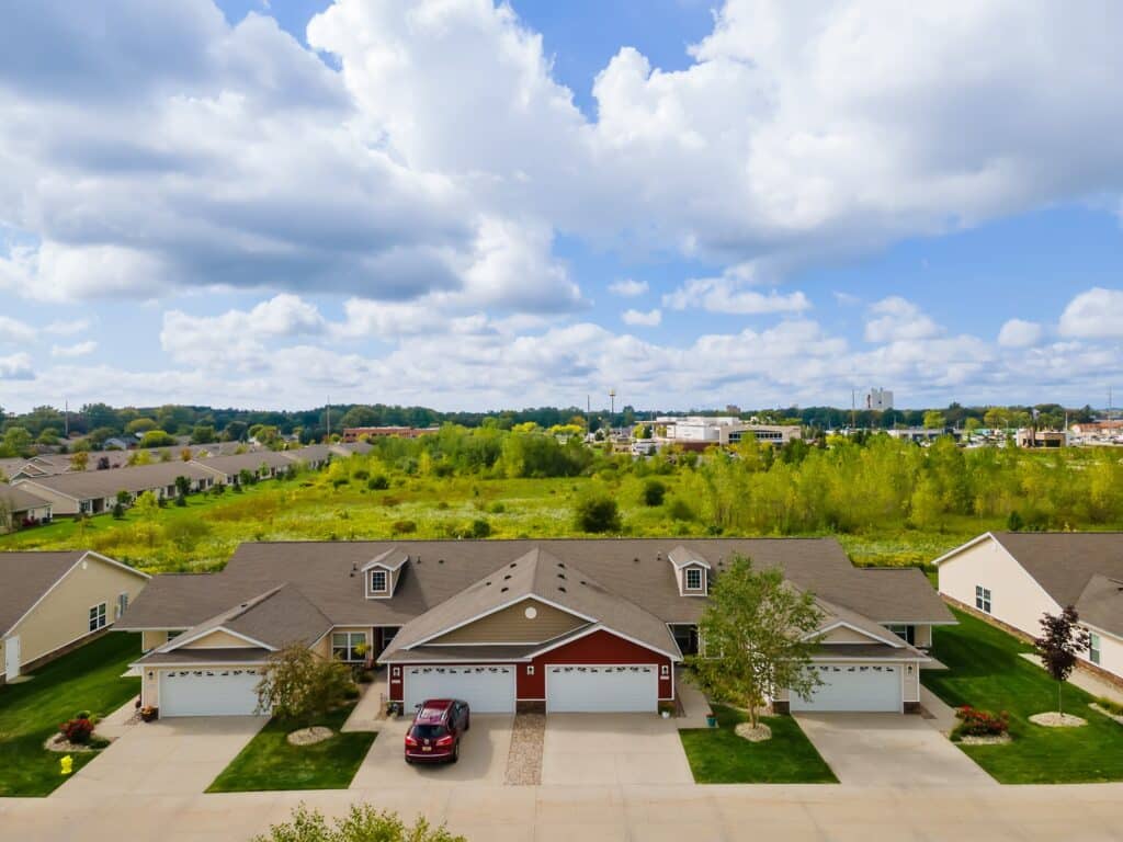 A row of single-story townhomes with garages and driveways, set in front of a grassy field under a partly cloudy sky.