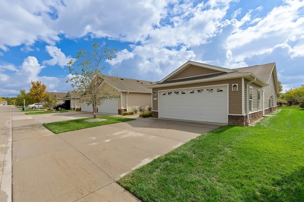 A row of suburban single-story houses with double garages, driveways, green lawns, and a partly cloudy sky overhead.