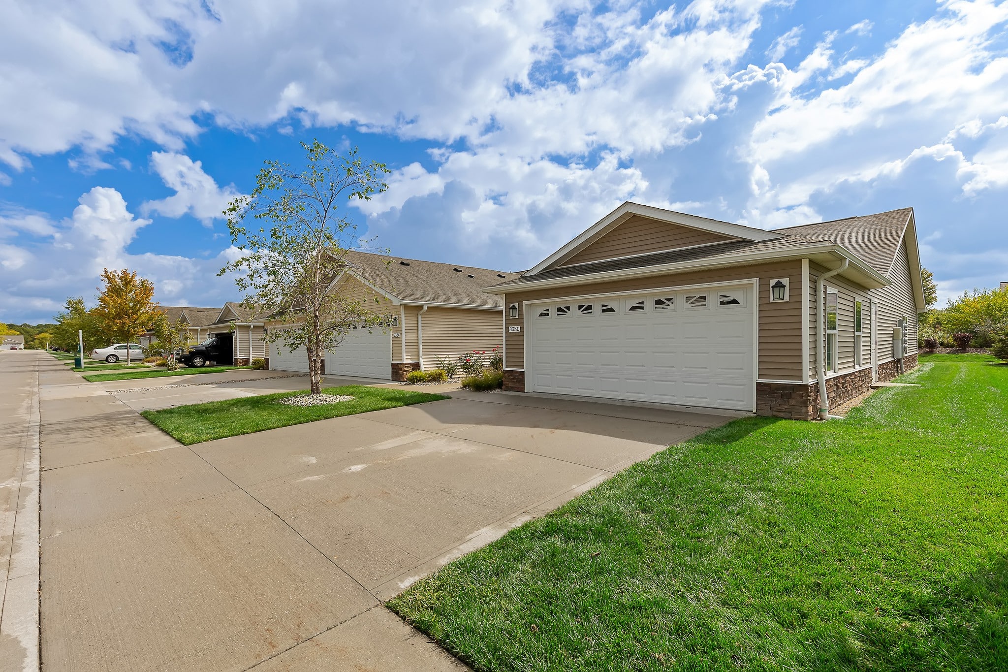A row of suburban single-story houses with double garages, driveways, green lawns, and a partly cloudy sky overhead.