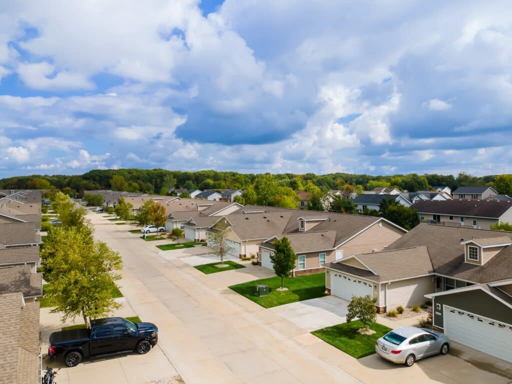 Aerial view of a suburban neighborhood with rows of single-family houses, green lawns, parked cars, and trees under a partly cloudy sky.