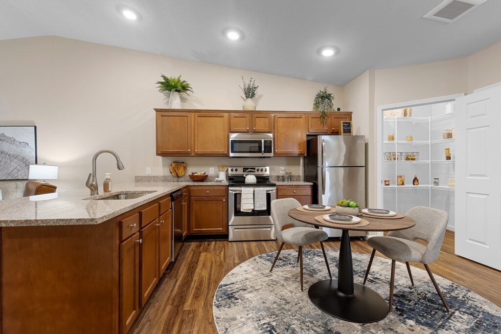 Modern kitchen with wooden cabinets, stainless steel appliances, a round dining table with three chairs, and an open pantry stocked with food items.