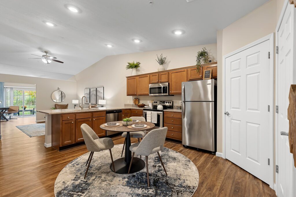 Modern open kitchen with wood cabinets, stainless steel appliances, a round dining table with two chairs, and a rug. Adjacent living and dining areas are visible in the background.